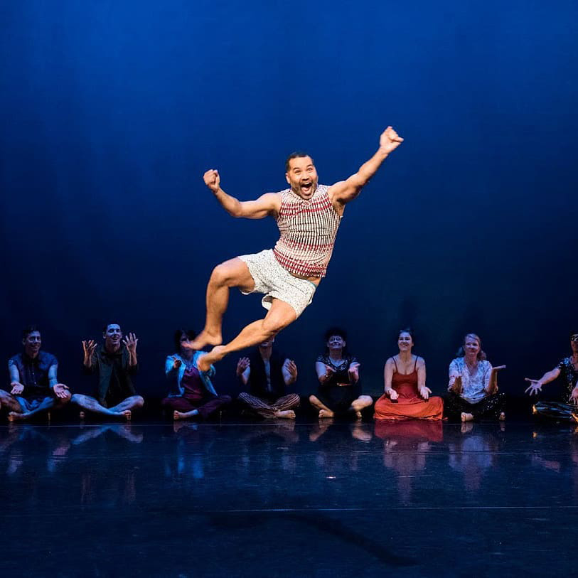 On stage, a performer is jumping in the air with a big open smile on his face. While jumping, his legs are slightly off to the side where he touches his heels together, and his arms are stretched out to both sides, as if he's punching the air. Behind him there are 8 people are sitting along the stage, smiling and holding their hands out in front of them.