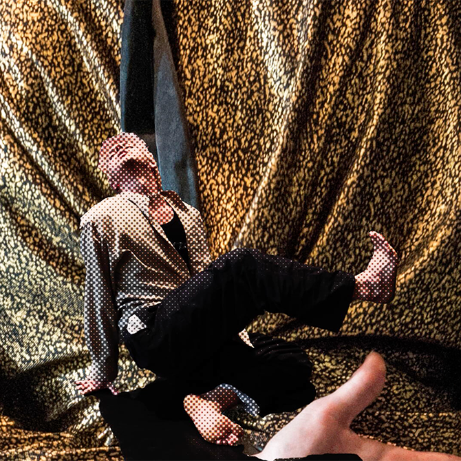 A cut-out of a dancer propping themselves up from the floor while lifting and crossing their leg over to the other side of them. The cut out is in front of a background of gold, shimmering fabric from a costume.