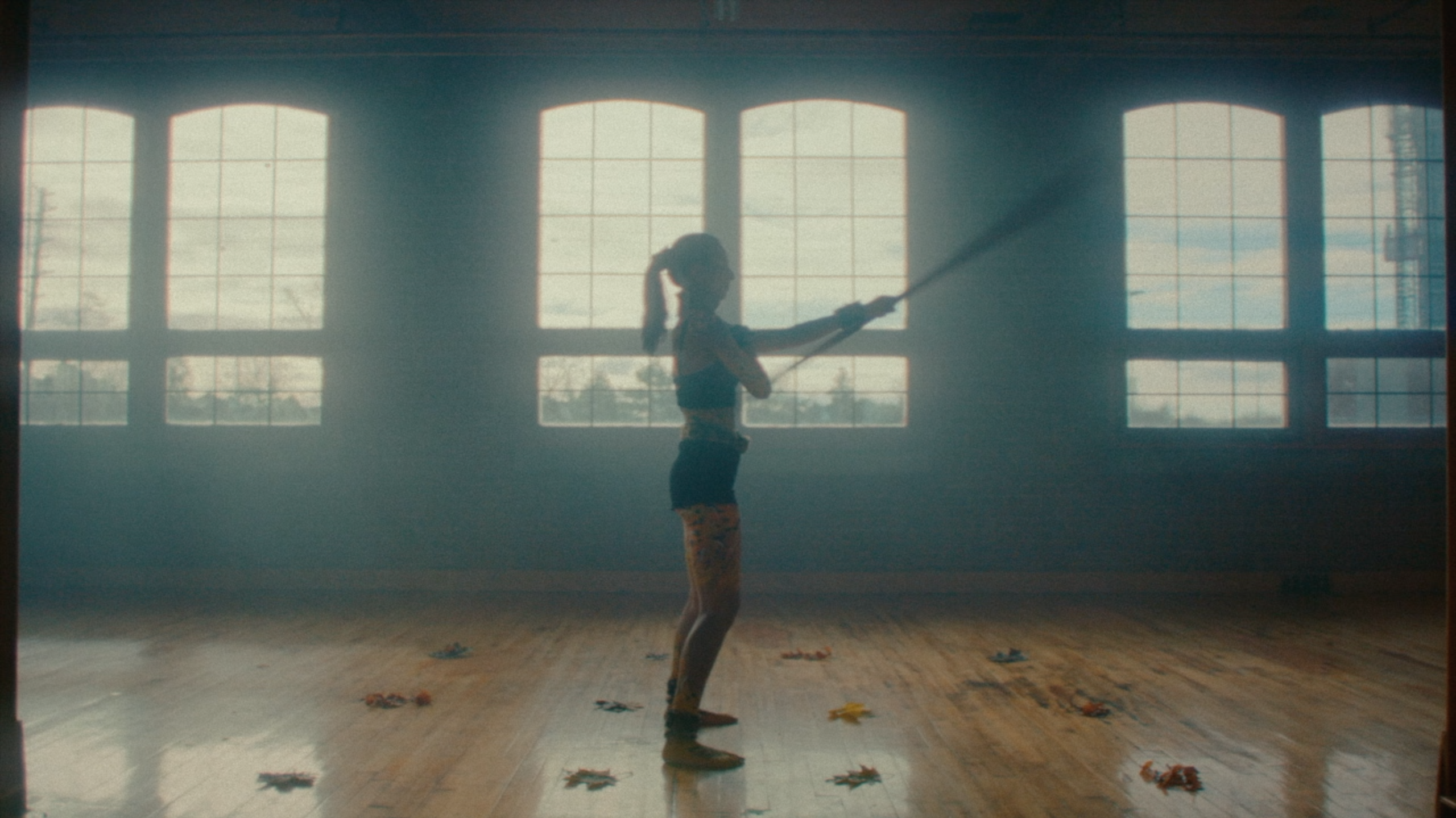 A dancer is standing in an open space with large windows behind them. The artist is facing the right side and moving a long wooden dowel out in front of her.