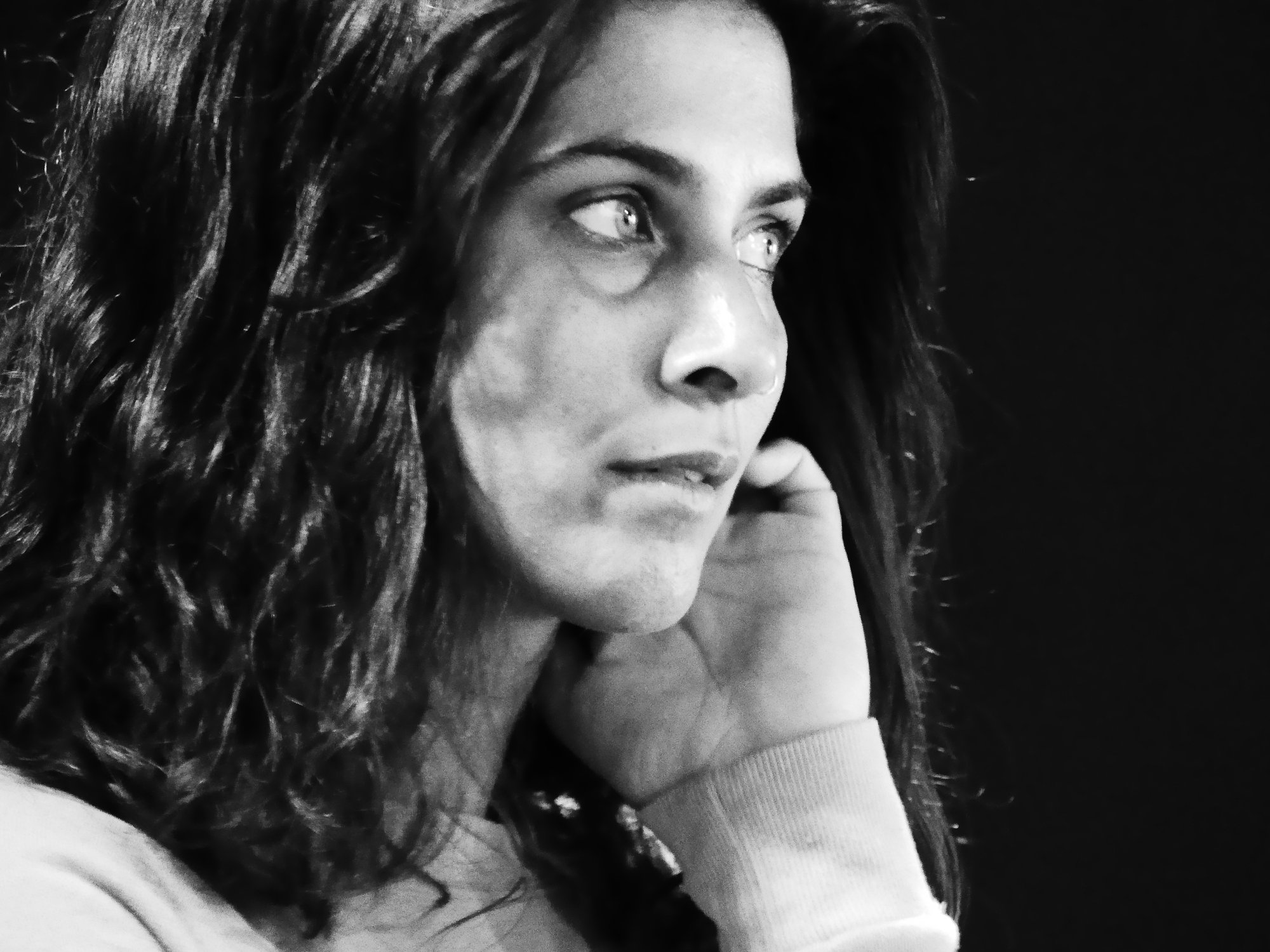 A black and white headshot in side profile of a brown femme with light eyes, dark wavy hair, nosering, holding hand to cheek and gazing into the distance.