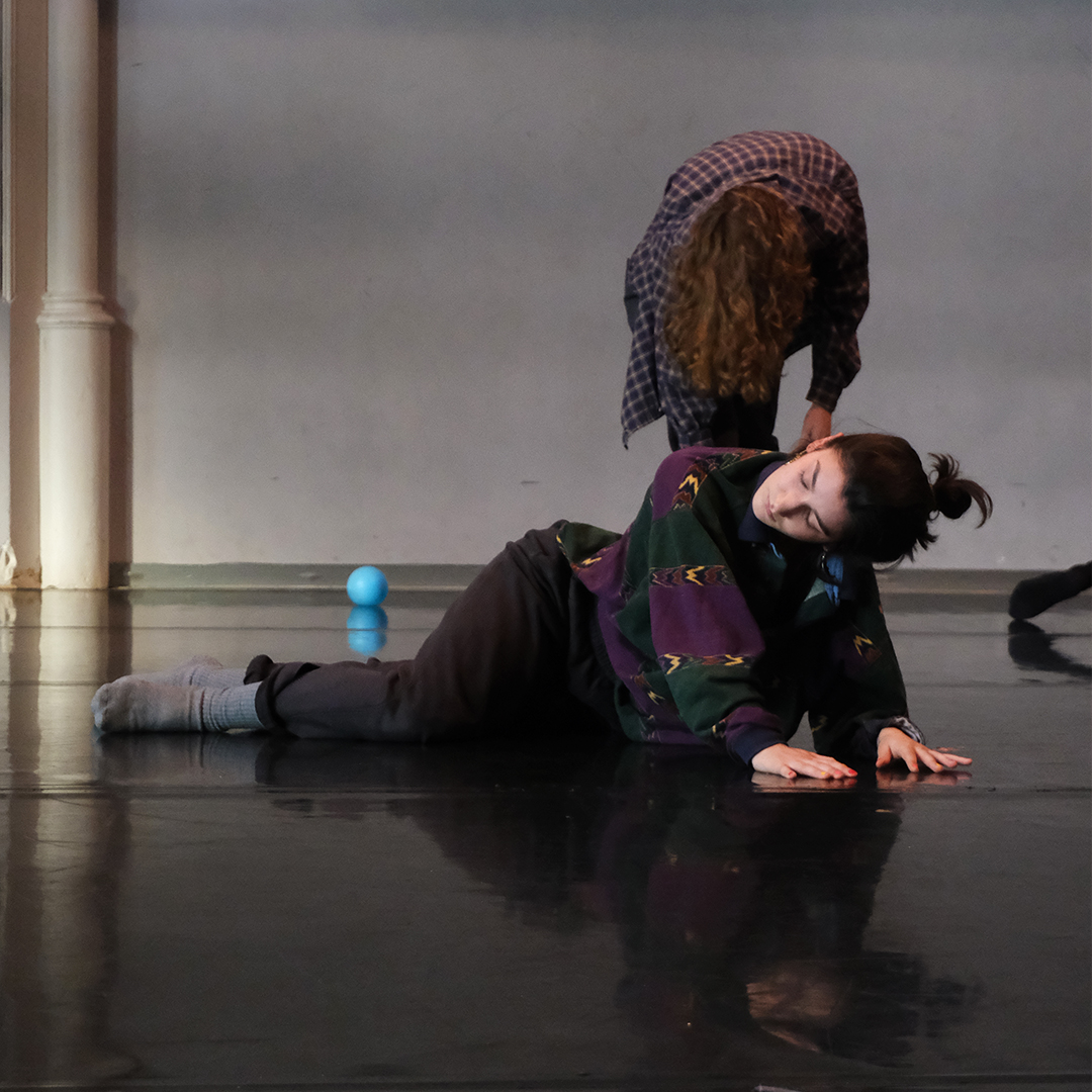 A dance artist is moving while laying on the floor of a studio. She is pushing her upper body up with her arms while her legs stay relaxed across the floor.