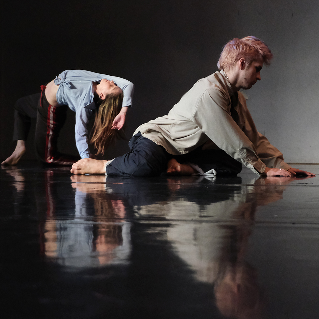 Two dancers are moving on the floor of a studio, and you can see their reflections shimmer across the floor. The dancer on the left is further back and has lifted herself into a backwards bridge, holding herself up with one arm and the other arm partially covering her face. The other dancer is closer to the camera, sitting on his shins while putting his hands on the ground in front of him and looking down.