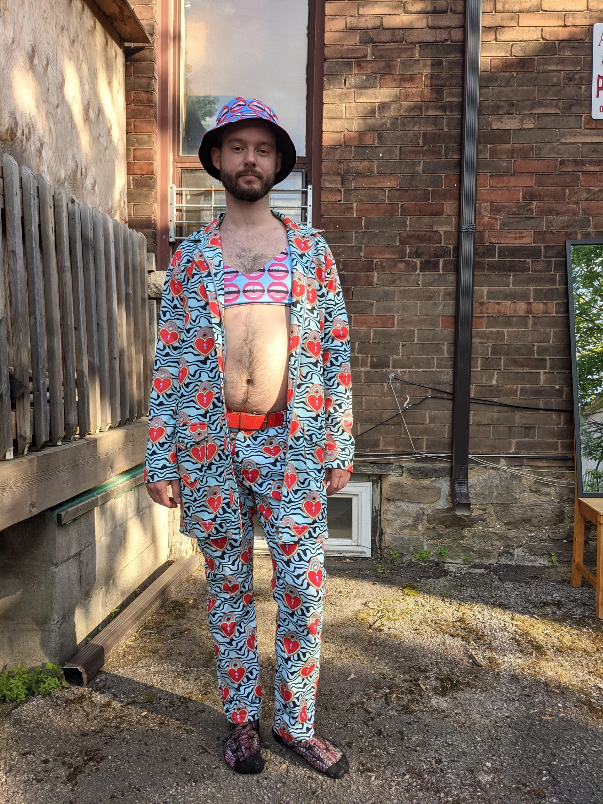 Jordan, a tall thin white gay man with a beard, is standing in a backyard, looking directly at the camera. He is wearing head-to-toe flashy, brightly coloured pop-art print (designed by Hayley Elasaeeser), including a bucket hat and bikini top with a matching mouthy print, and a jacket and pants with matching psychedelic green print. He is in bare feet, standing on pavement with a brick wall behind him.