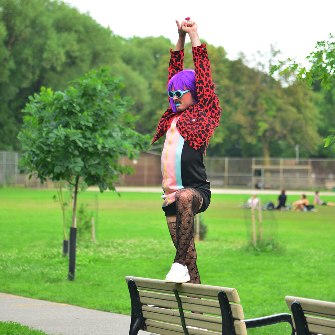 Jord Camp performing in an outdoor public park. She is standing on a park bench with one foot up on the edge, arms to the sky. It is a bright, sunny summer day and there is green grass and trees surrounding the performer. Jord is wearing a purple bob wig, blue sunglasses, blue lipstick, and a red cropped jean jacket over a colour-blocked dress with stripes of light pink, baby blue and black. She is wearing fishnet tights.