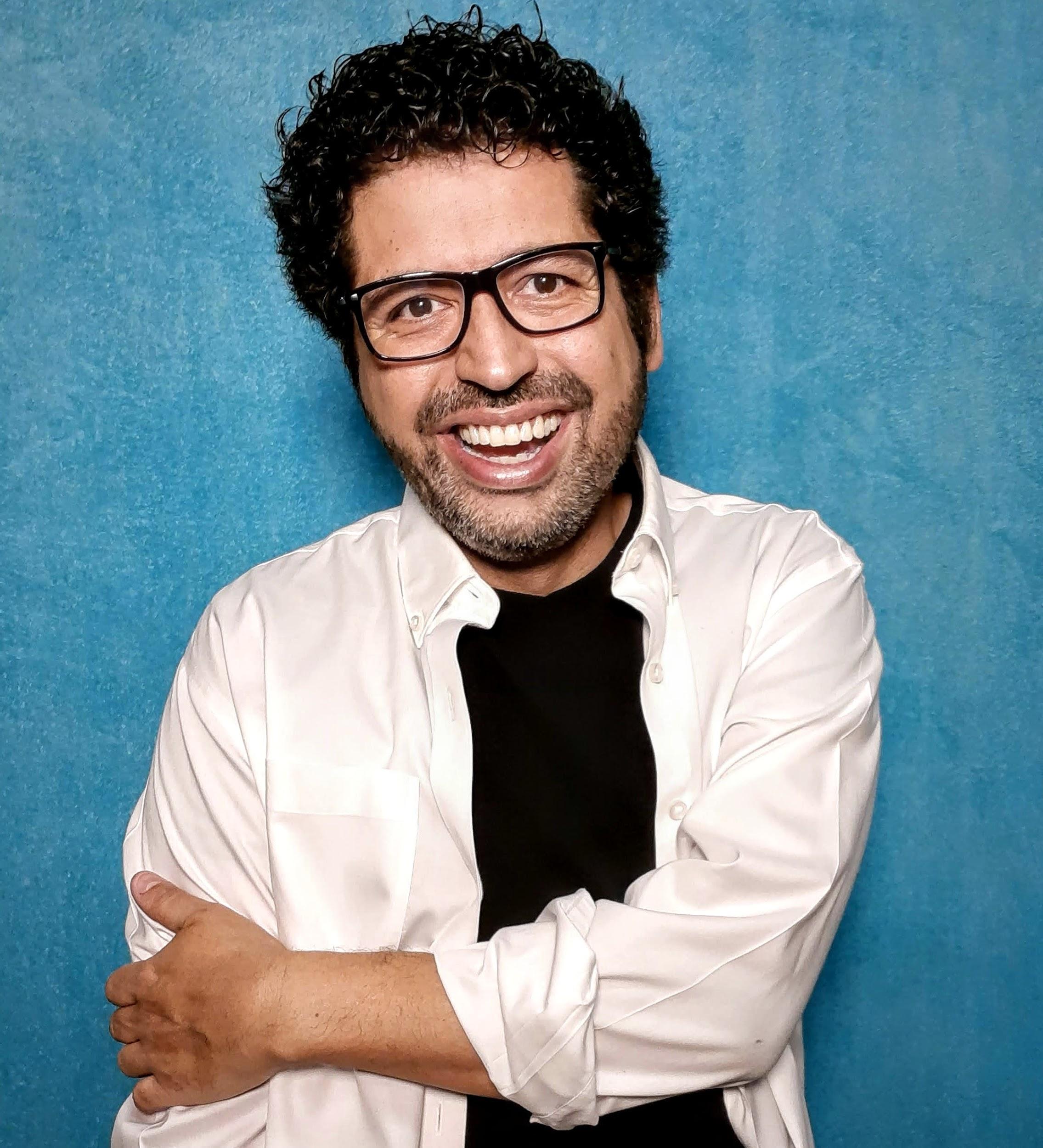 Portrait of Juan smiling at the camera in front of a blue background, wearing black glasses and a black tee with an open white collared shirt over top.
