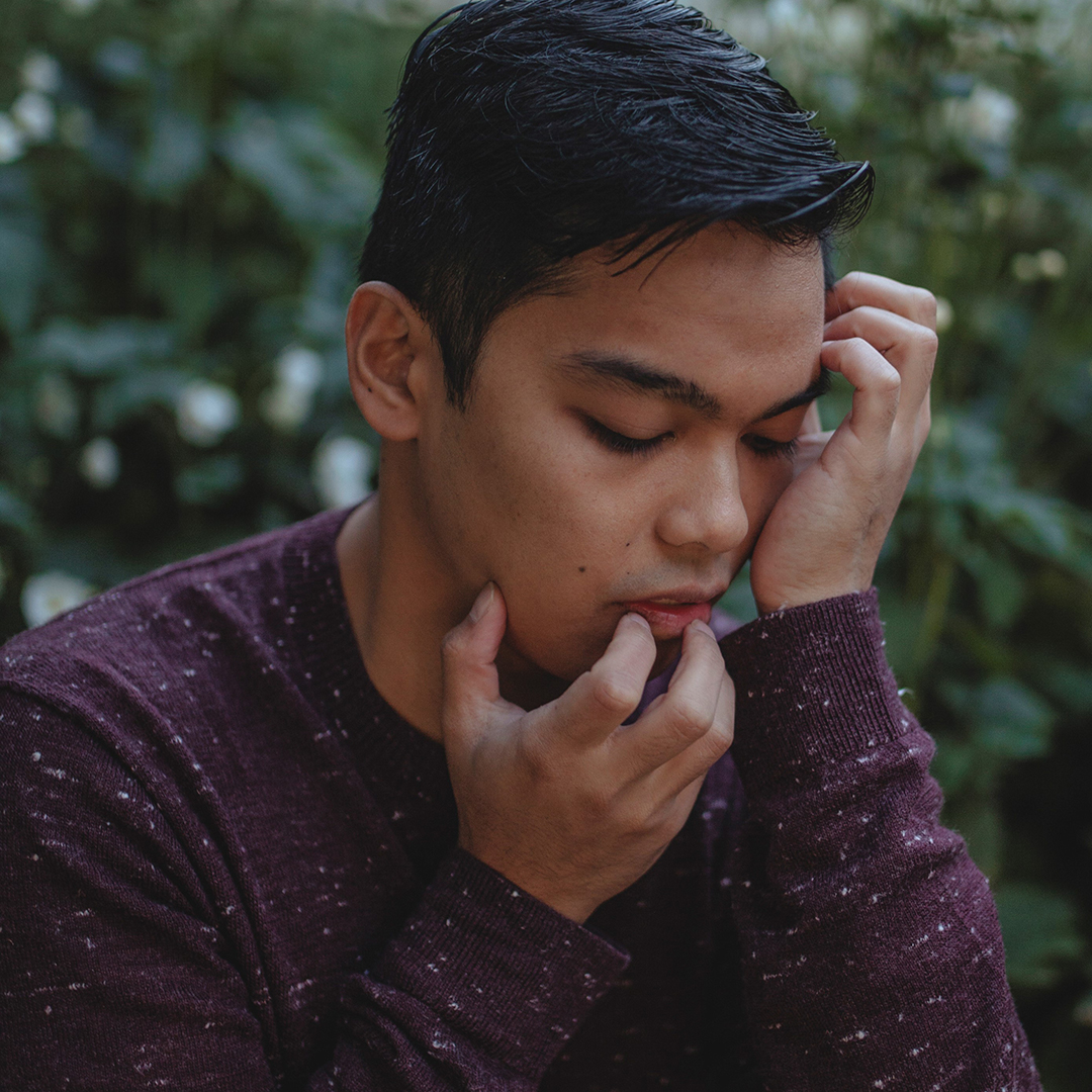 A side profile of Miggy, a brown-skinned man wearing a long-sleeved maroon shirt speckled with grey. He is in front of a bush, looking down with eyes closed, as his fingers gently crawl over his face.