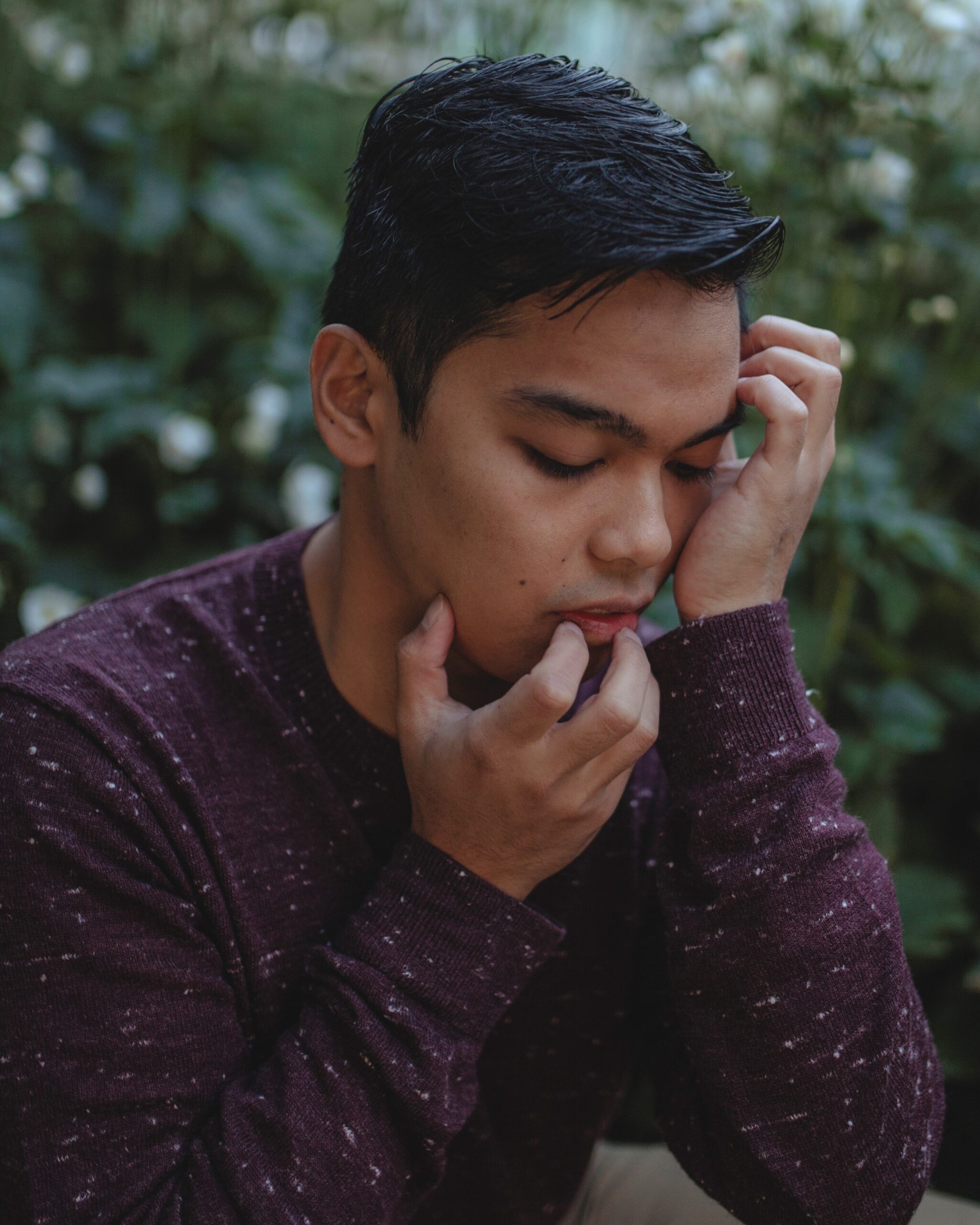 A side profile of Miggy, a brown-skinned man wearing a long-sleeved maroon shirt speckled with grey. He is in front of a bush, looking down with eyes closed, as his fingers gently crawl over his face.
