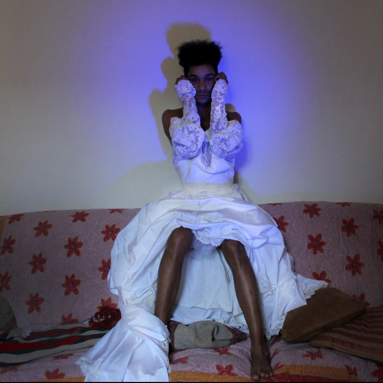 Shadé is sitting on a couch wearing a white ball gown and satin gloves, holding their forearms up on both sides of their face, while a purple spotlight glows on their face.