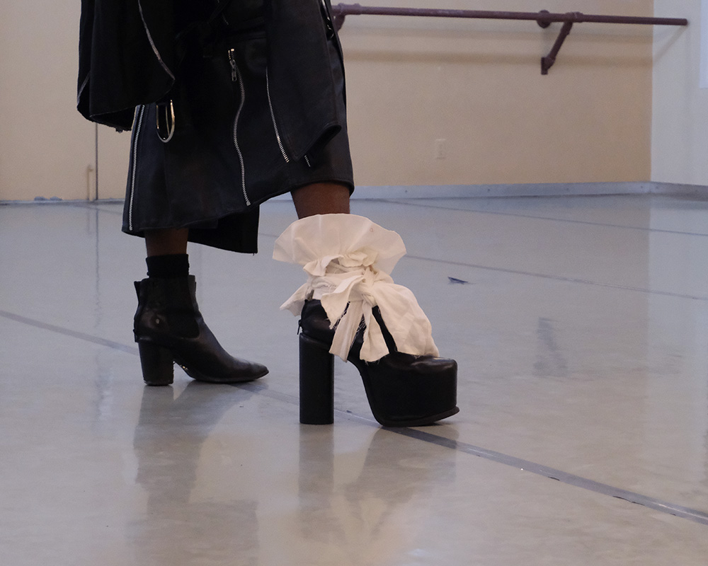 A close-of the high heels that a dancer is wearing. The heels are at two different heights, with the right heel having white fabric tied around the ankle in a knot.