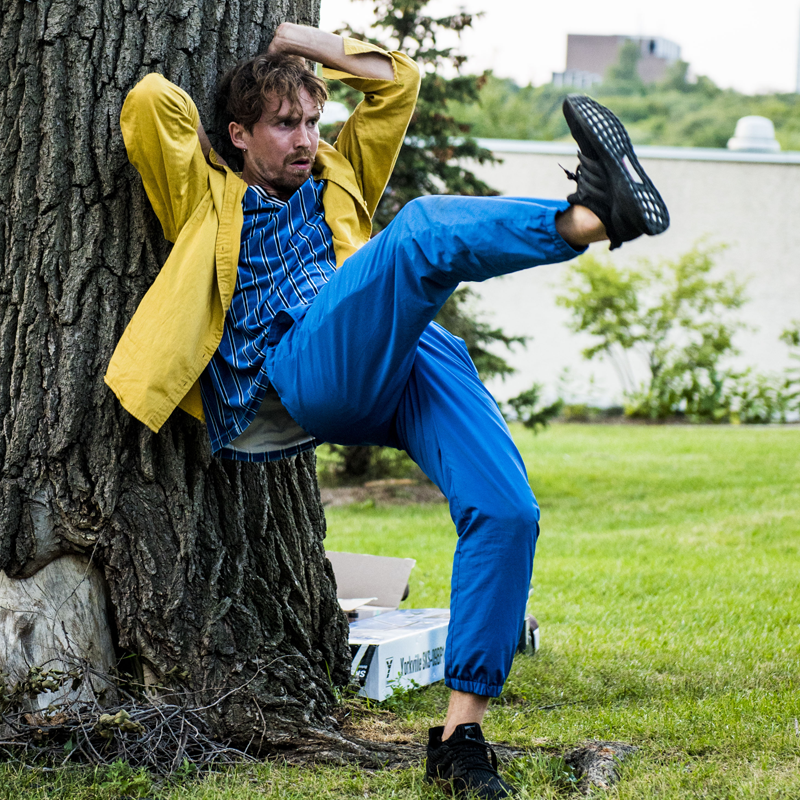 A photo of Lukas Malkowski dancing. He's leaning against a tree, hands overhead, balanced on one leg while the other kicks high and outward.