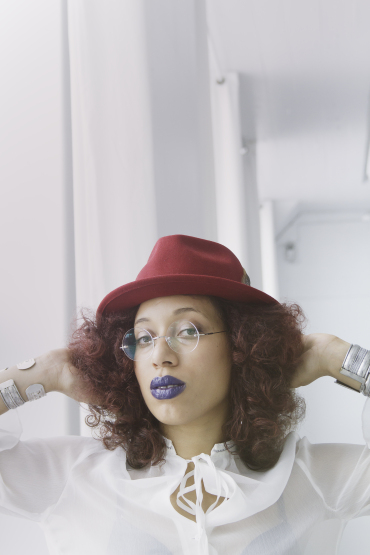 A photo of Sam I Am Montolla. She is a medium-light skinned person with cherry red curly hair. She's wearing a red hat, round glasses and has purple lipstick. Her hands are in her hair as she stairs at the camera.