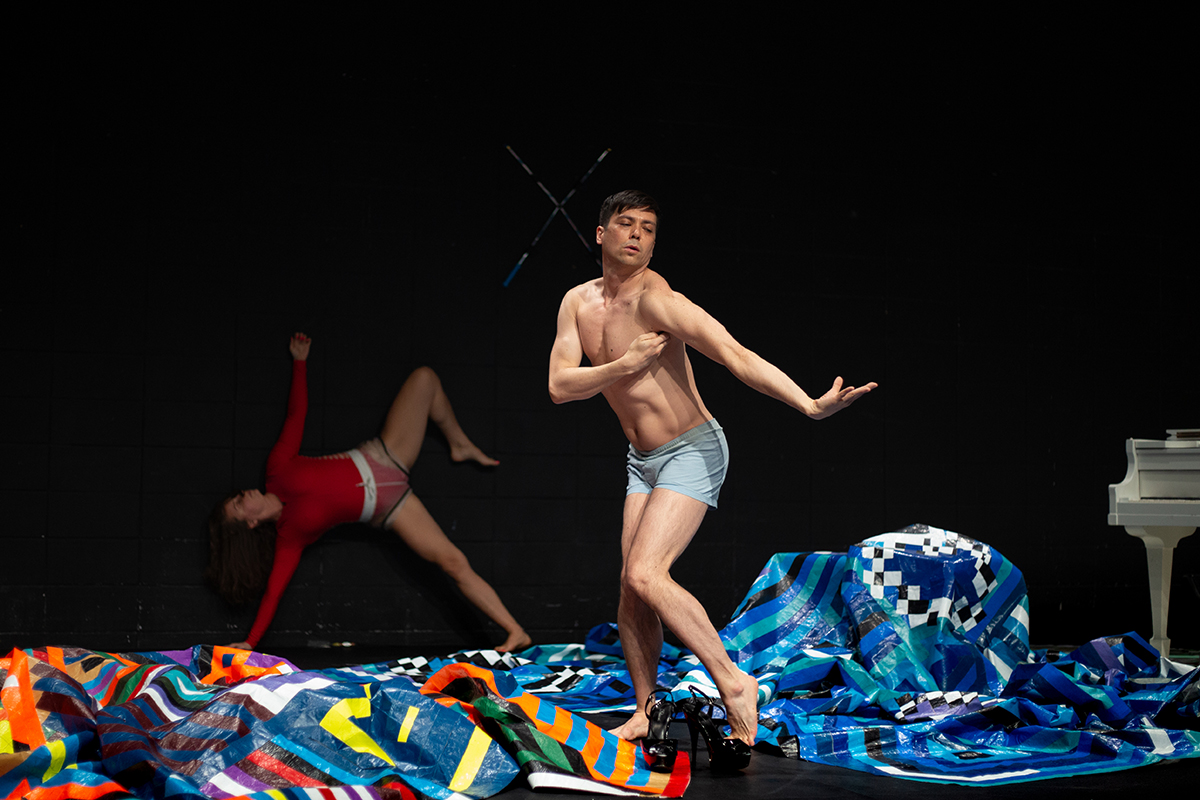 Andrew Tay is in the centre, wearing light blue briefs and dancing on stage with bright, multi-coloured tarps covering most of the floor. Andrew is standing with his body slightly turned to the left, but he is looking back over his shoulder while stretching the arm out behind him and bending his hand back. At the back of the photo on the left, there is another dancer in a bright red bodysuit, holding themselves up from the ground with one arm and leg, while reaching out their other arm up above them and bending their other leg in the air.