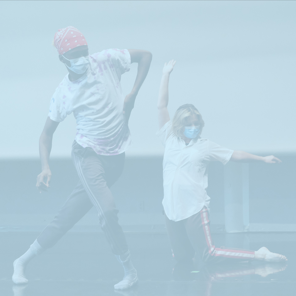 Two dancers are moving in the studio. One is kneeling, arms raised overhead. The other dancer side-steps, arms swinging.