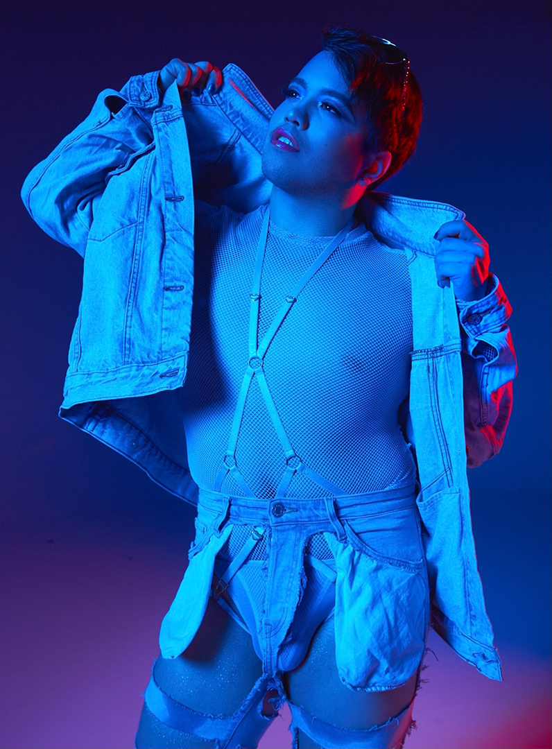 In front of a deep gradient blue background, Tygr Willy is standing in a blue glow, wearing a mesh body suit and denim shorts that were cut out except for the frame and its pockets. They are wearing an open denim jacket over top.