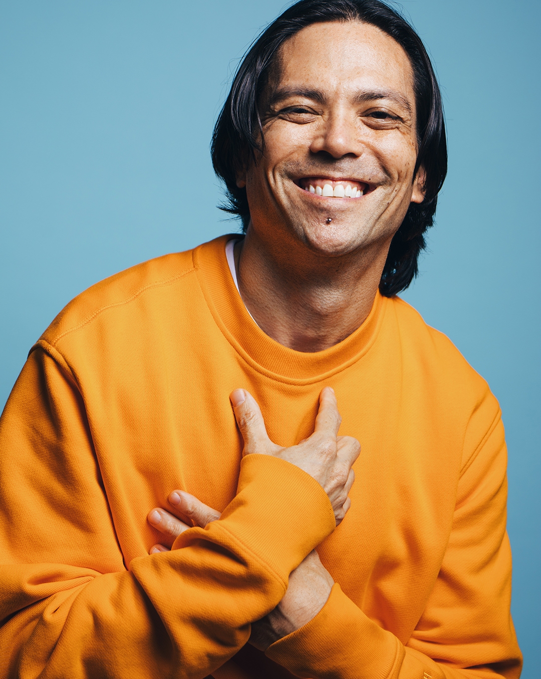 Portrait of Andrew Tay in front of a baby blue background while wearing a vibrant orange sweatshirt. While smiling big at the camera, he gently crosses his hands over one another on his chest.