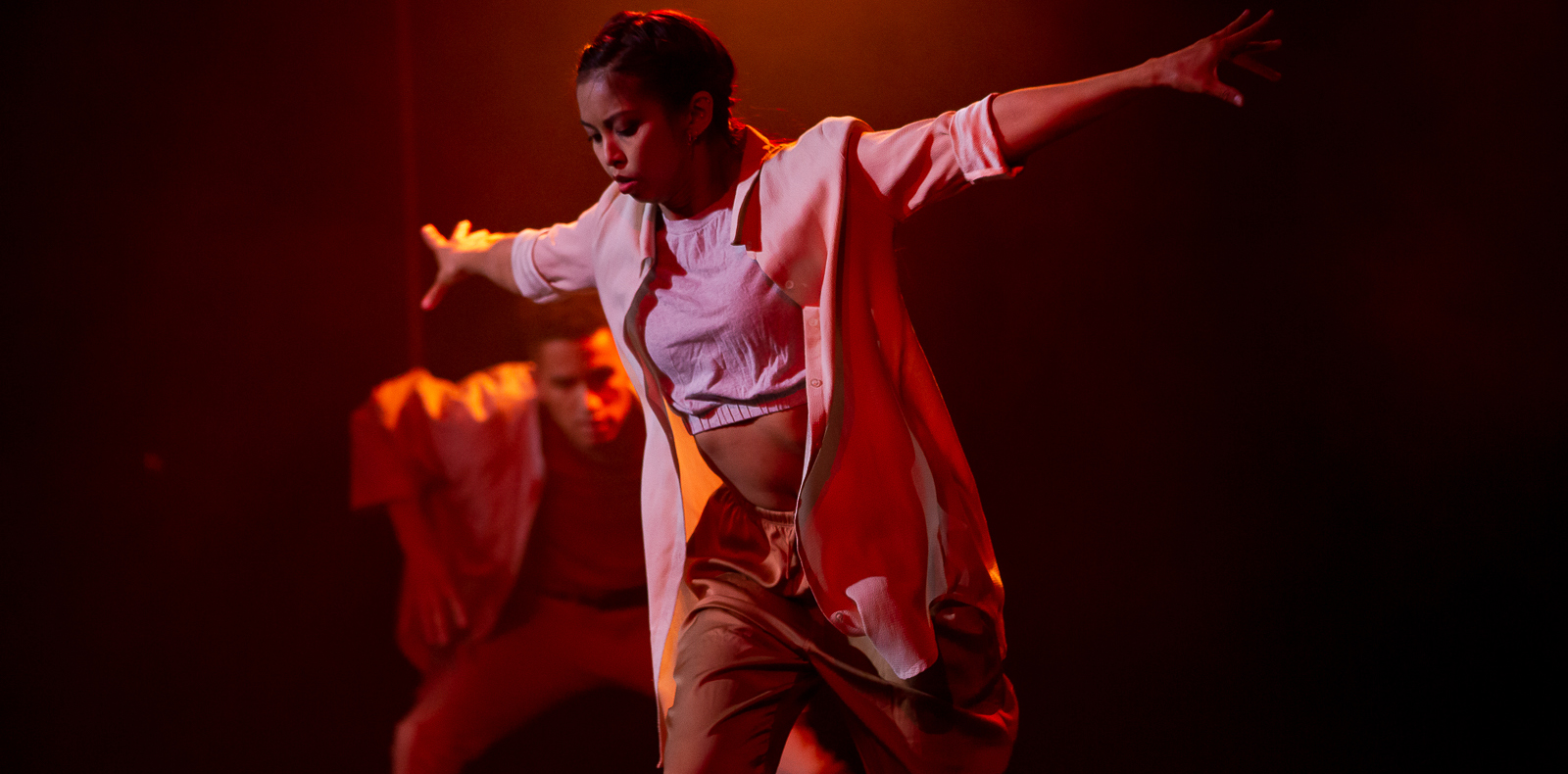 Two performers are doing a duet on an orange and red-lit stage. One of them is near the back, slightly squatting as he watches the other dancer stick her arms straight out from her sides at shoulder height while she stands on one leg and looks down.