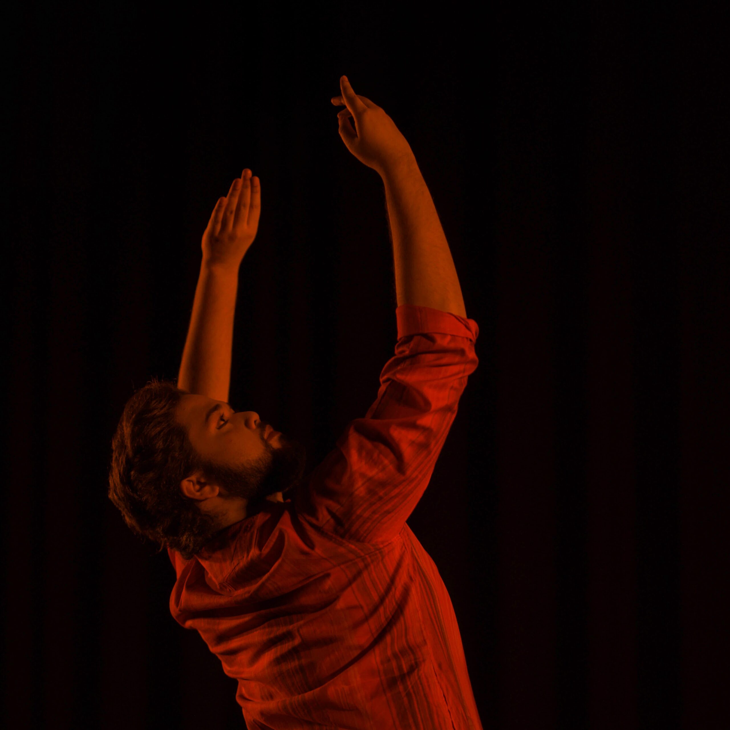 Dancing in front of a black background, Tanveer Alam is pivoted towards the back while looking up to the right said and curving both of his arms over his head