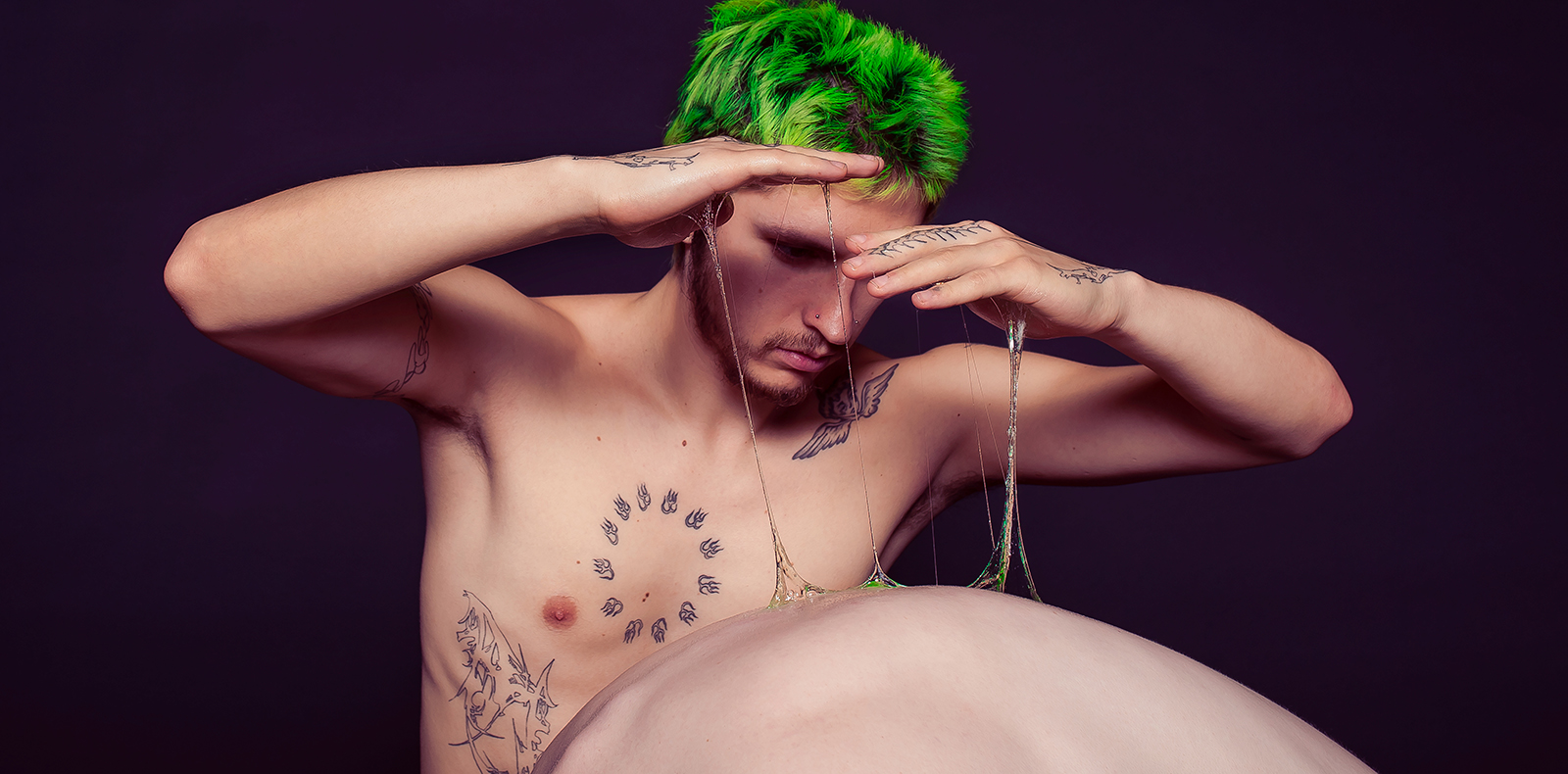 A dance artist with bright green hair is shirtless, playing with gooey, gel-like liquid and stretching it up from another dancer's back, whose face you cannot see and they are contracting their back.