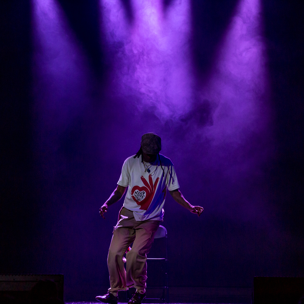 A dancer is performing on stage underneath purple, hazy spotlights. He is standing with his head tilted down, twisting his legs and feet a little to the left side.
