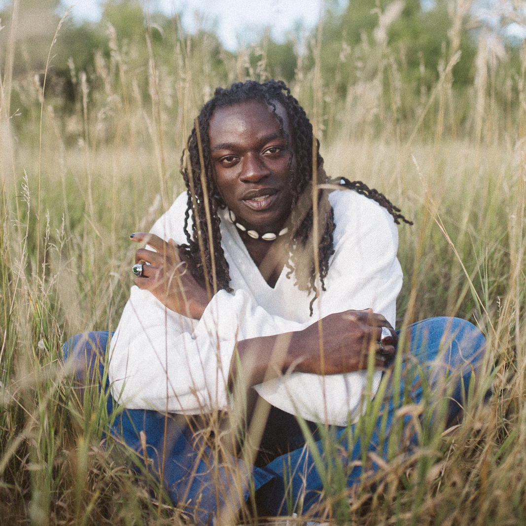 Kwasi Obeng is sitting cross-legged in an open field of golden ornamental grass, smiling at the camera. He wears a white long-sleeved shirt and blue jeans.