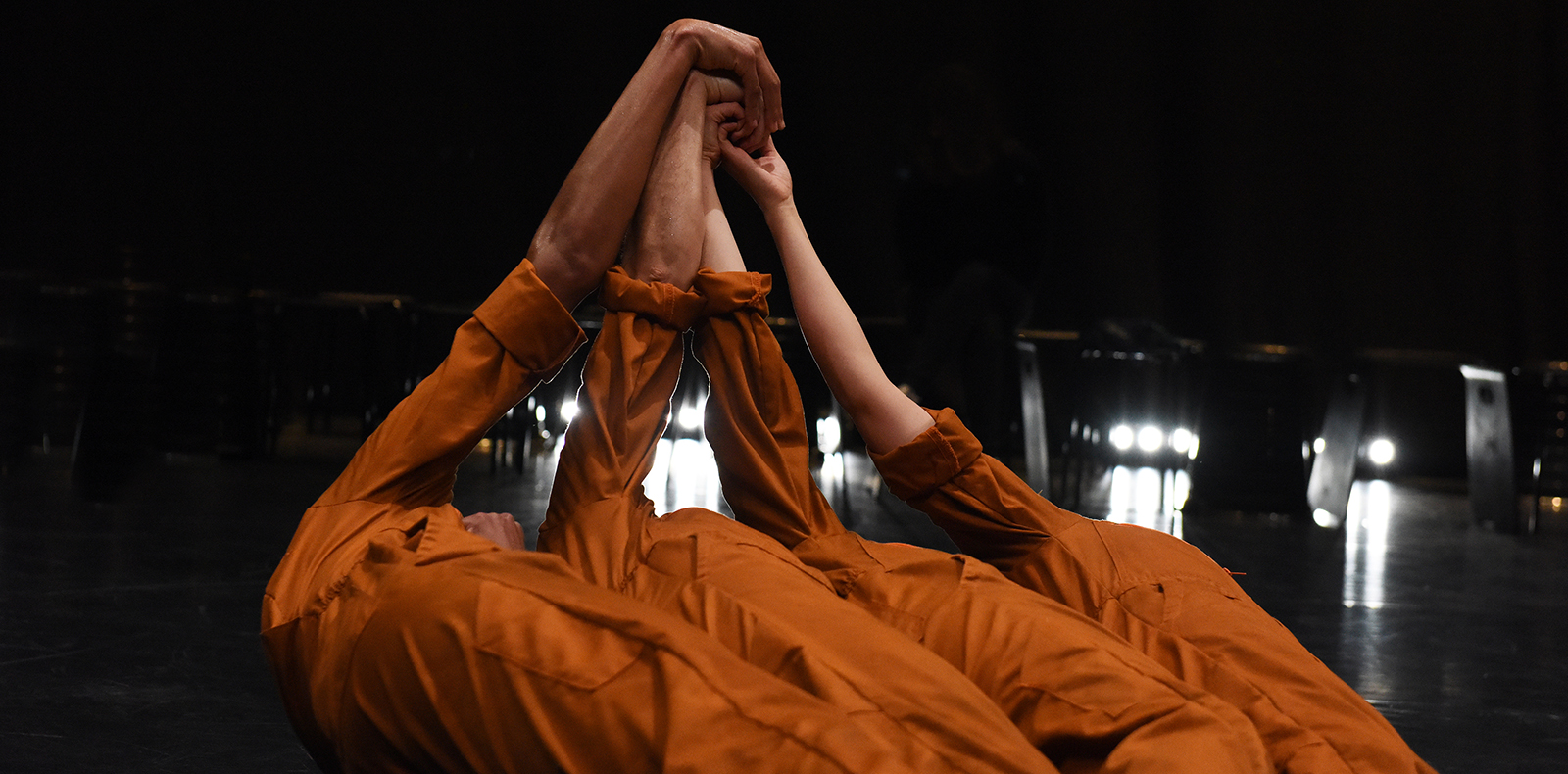 Four dancers in burnt-orange jumpsuits are leaning back alongside each other and you can't see their faces. They are each extending out an arm above them towards the centre to clasp their hands altogether.