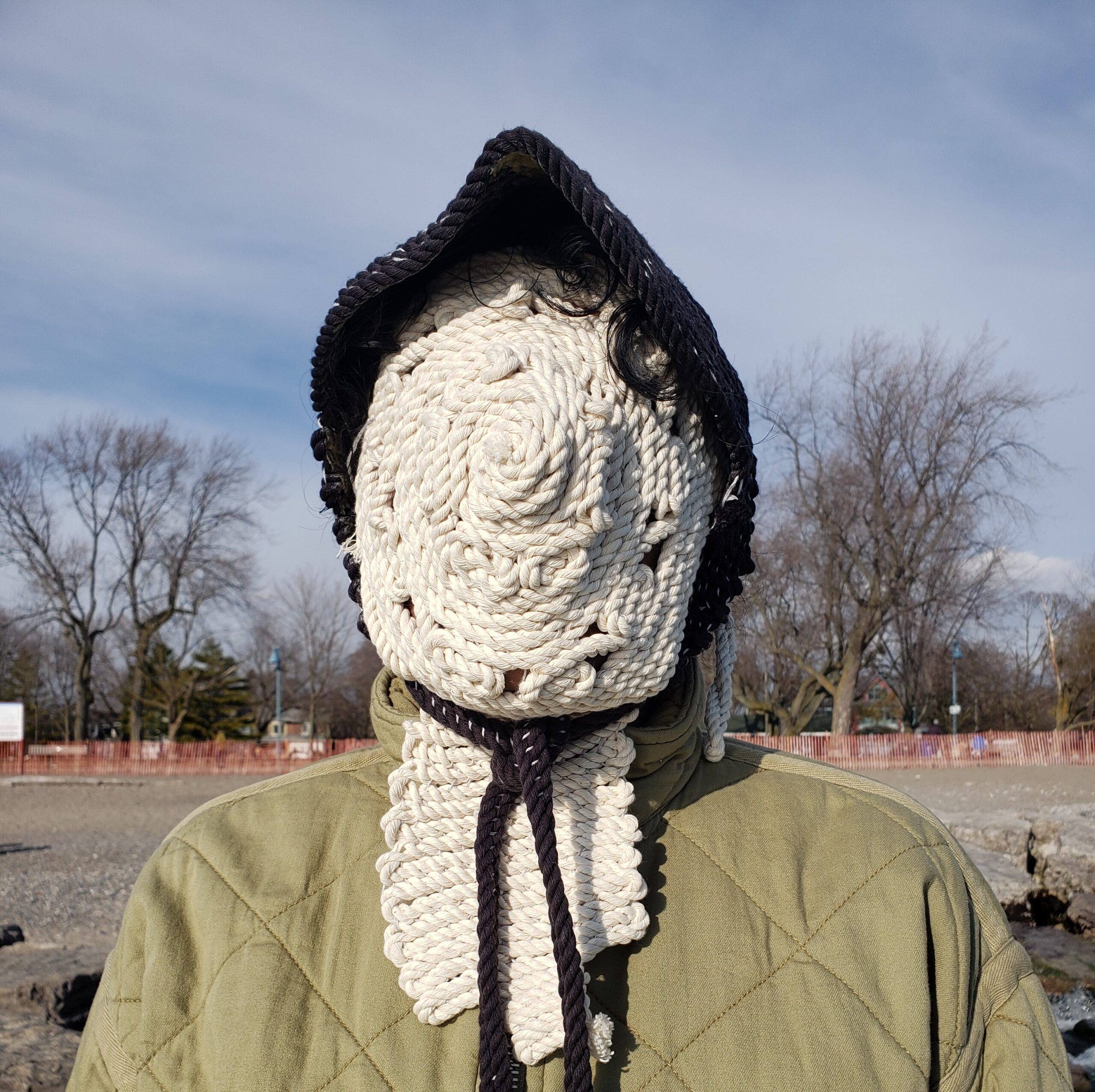 Akash is outdoors, wearing a green sweater and their face and neck is completely covered by a textile mask of thick white rope that is stitched together in a circular pattern. They are wearing a black bonnet made of similar thick rope.