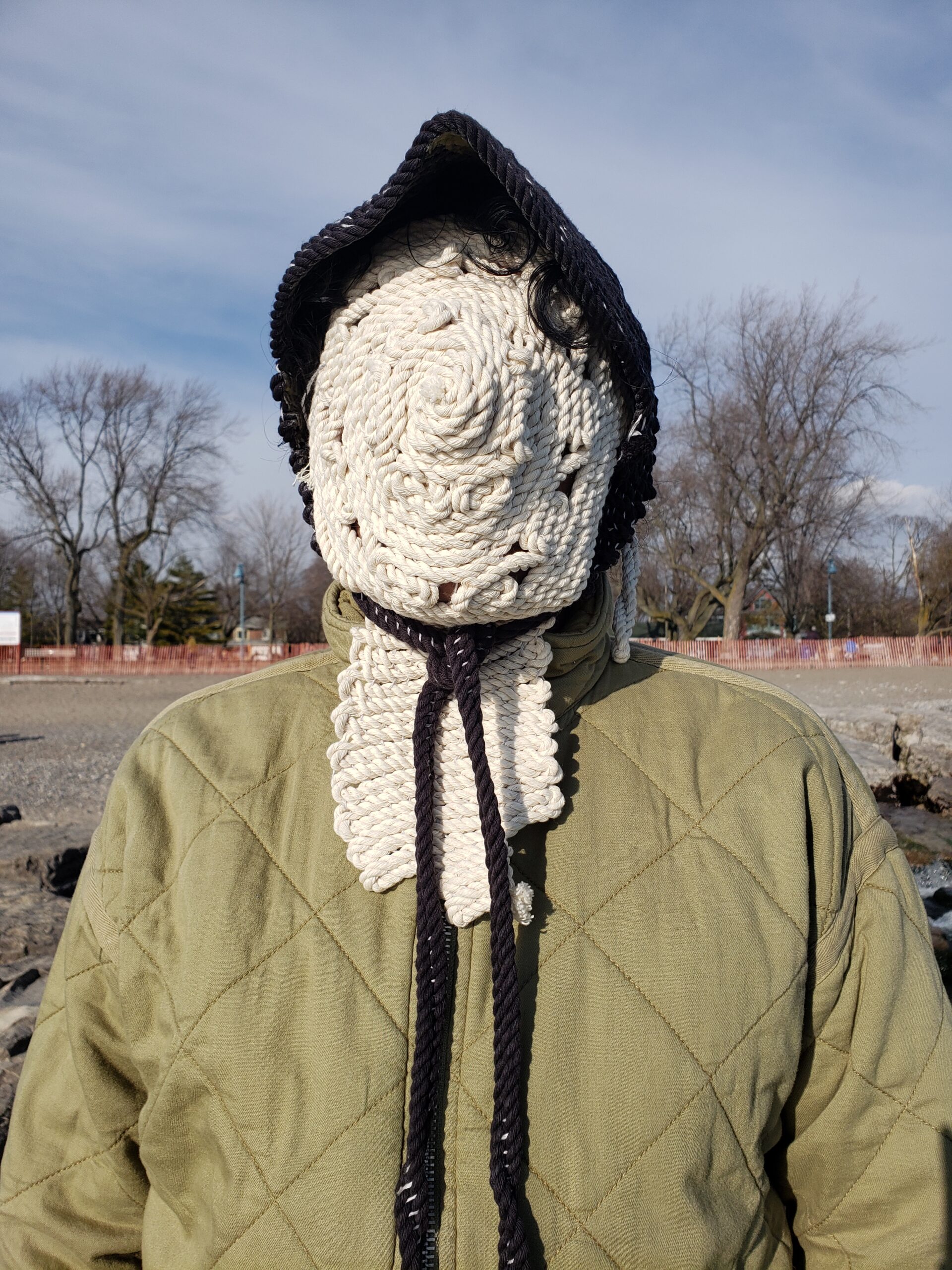 Akash is outdoors, wearing a green sweater and their face and neck is completely covered by a textile mask of thick white rope that is stitched together in a circular pattern. They are wearing a black bonnet made of similar thick rope.