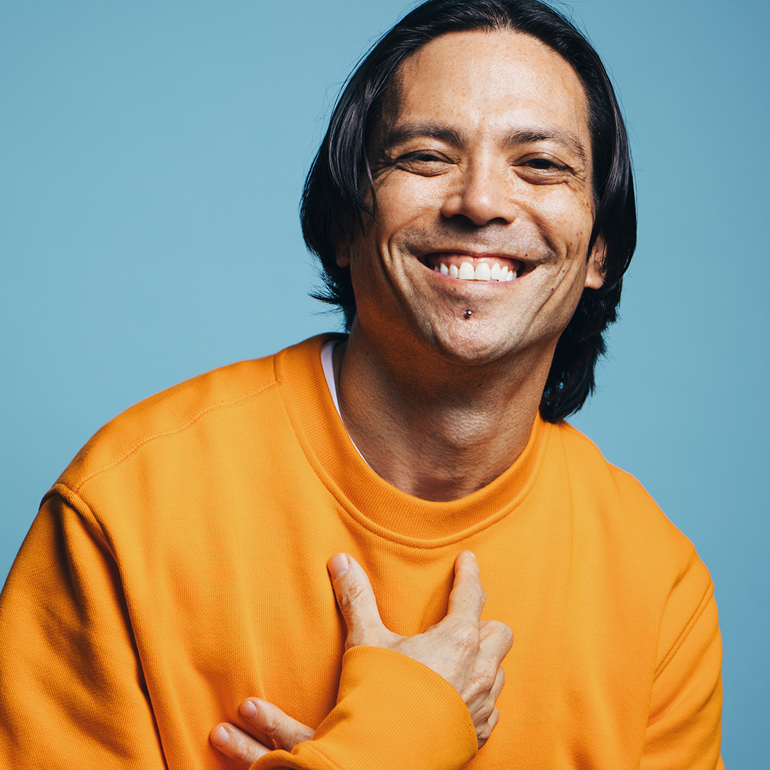 Portrait of Andrew Tay in front of a baby blue background while wearing a vibrant orange sweatshirt. While smiling big at the camera, he gently crosses his hands over one another on his chest.