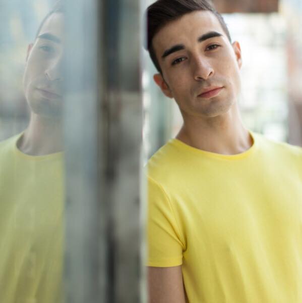 Headshot of Cody Berry in a bright yellow t-shirt, leaning his head and shoulder against a wall.