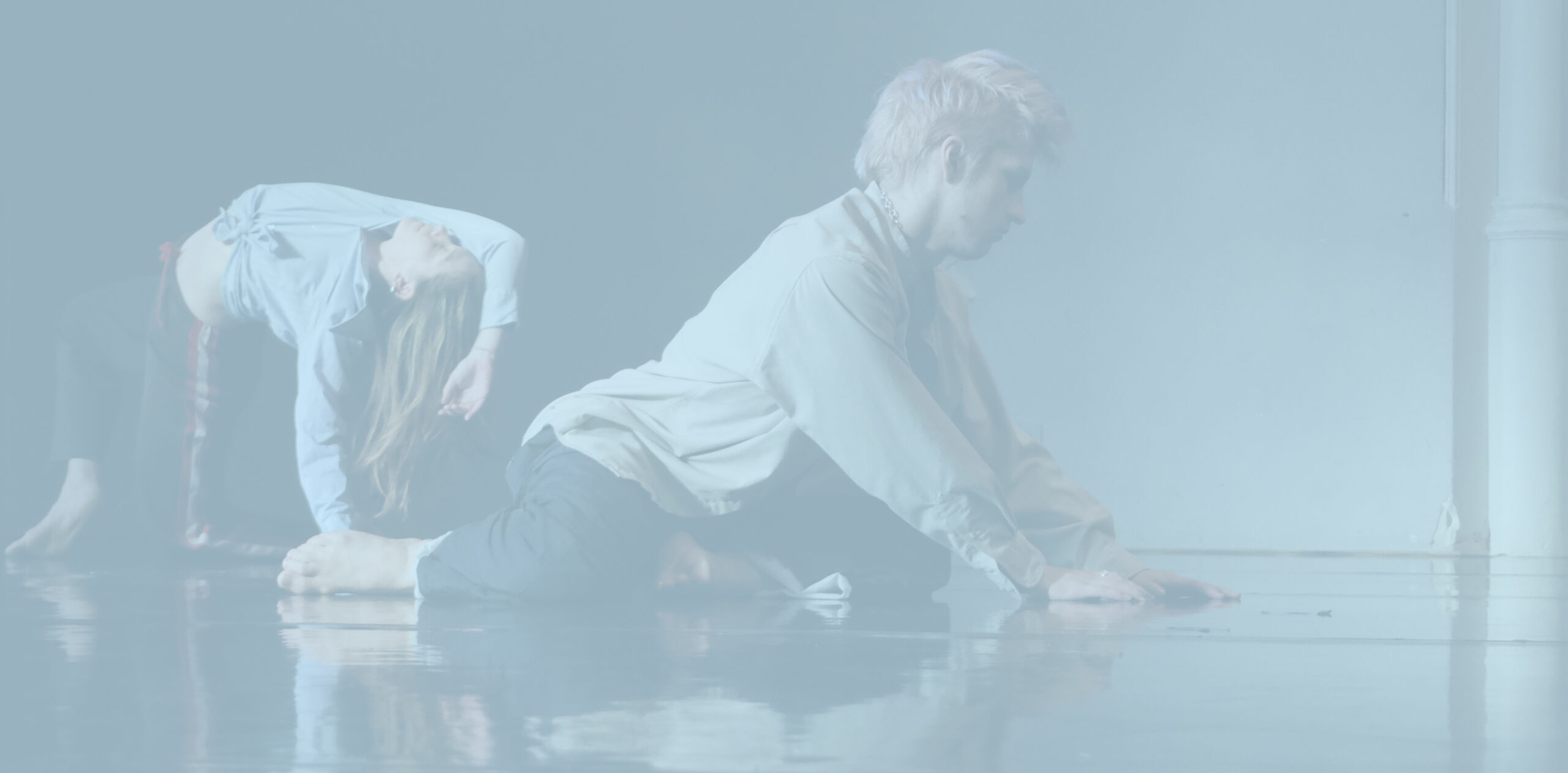 Two dancers are moving on the floor of a studio, and their reflections shimmer across the floor. The dancer on the left is further back and lifts herself into a backwards bridge, holding herself up with one arm and the other arm partially covering her face. The other dancer is closer to the camera, sitting on his shins while putting his hands on the ground in front of him and looking down. There is a pastel blue wash over the photo.