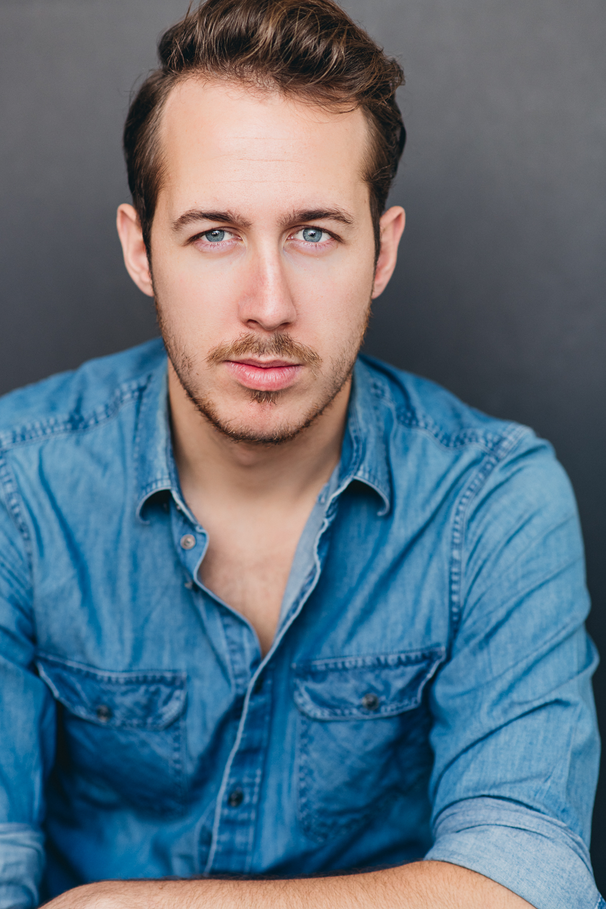 Portrait of Bryan in a blue denim buttoned-up shirt, in front of a grey background. They are looking directly at the camera.