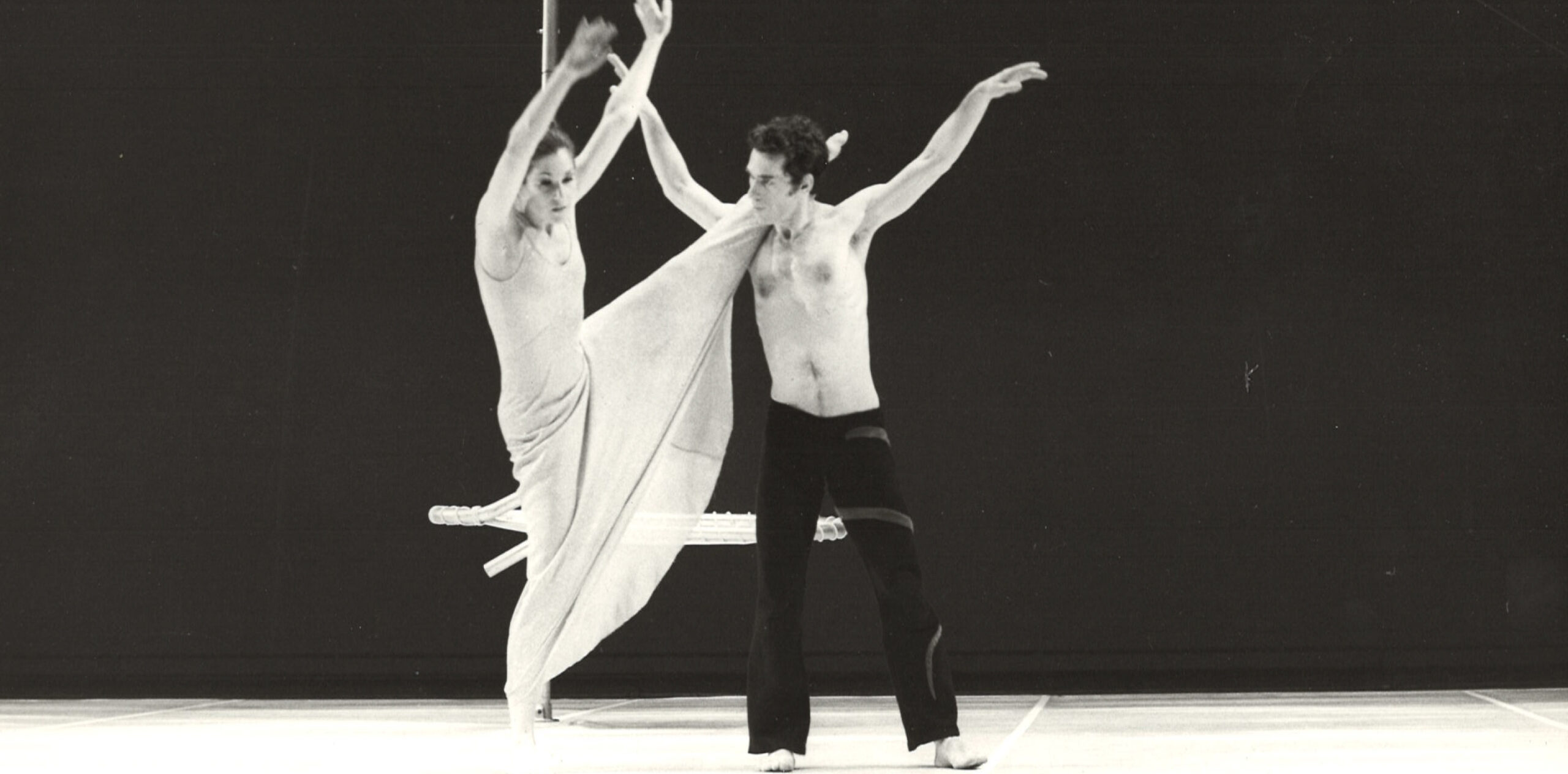 In an archive black-and-white photo, two dancers are performing on a stage. One dancer lifts her leg up straight and rests it on the other dancer's shoulder who is standing tall with his arms up towards the ceiling.