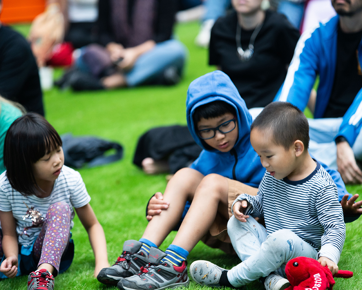 During a performance event, three kids are sitting on a large patch of grass with several other people behind them watching the performance.