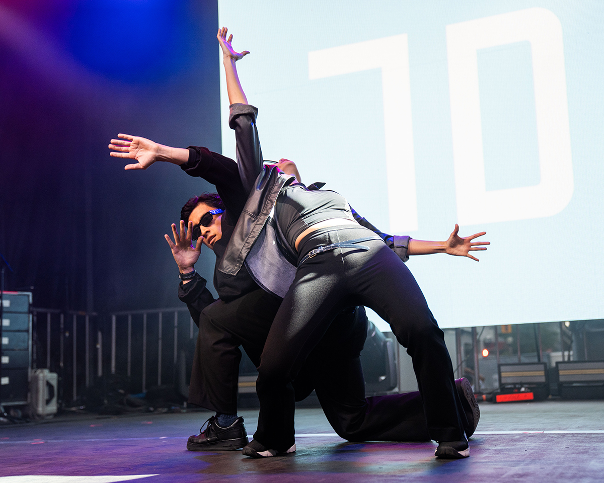 Two dance artists on performing on stage. Wearing all black, one artist is leaning backwards onto the other artist while they are each reaching out one of their arms in the air.