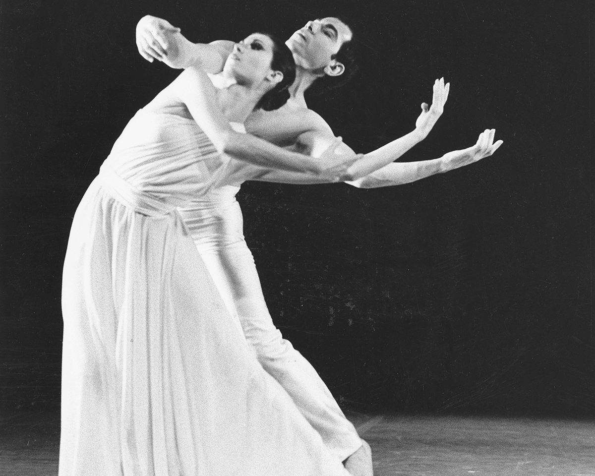 An archived black and white photo of two dancers performing. The dancer in the front is in a long and flowy light-coloured dress. Her hip sways to the left while she scoops her arms out to the right. The dancer behind her sways the same way while holding his arms out around her at shoulder height, tilting his head back, appearing as if his head sits gently right above her head.