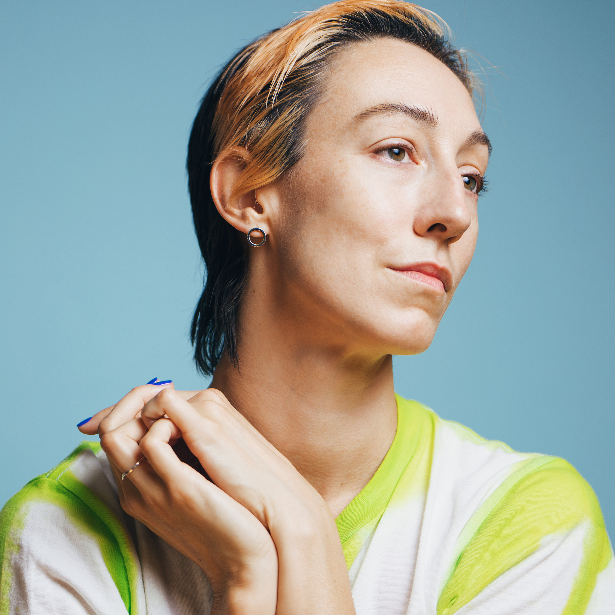 Portrait of Erin in front of a ice blue background, wearing a lime green and white stiped top. She clasps her hands and holds them closely to her right shoulder while looking towards the left.