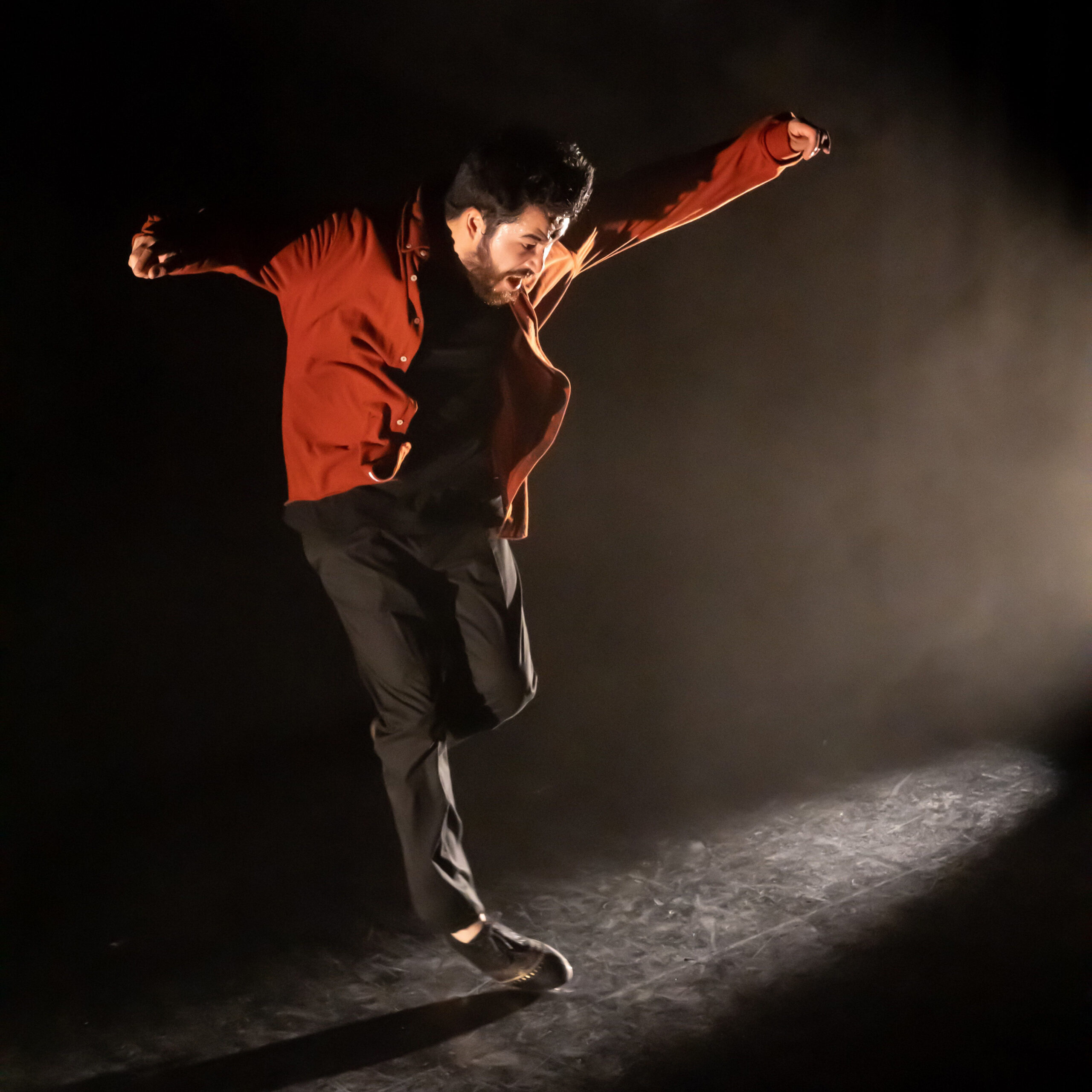 Johnny Morin is dancing in a red jack on a dark stage with a spotlight further away shining on his tap shoes. He holds his arms out from his sides while only the tip of one tap shoe is grazing across the floor.