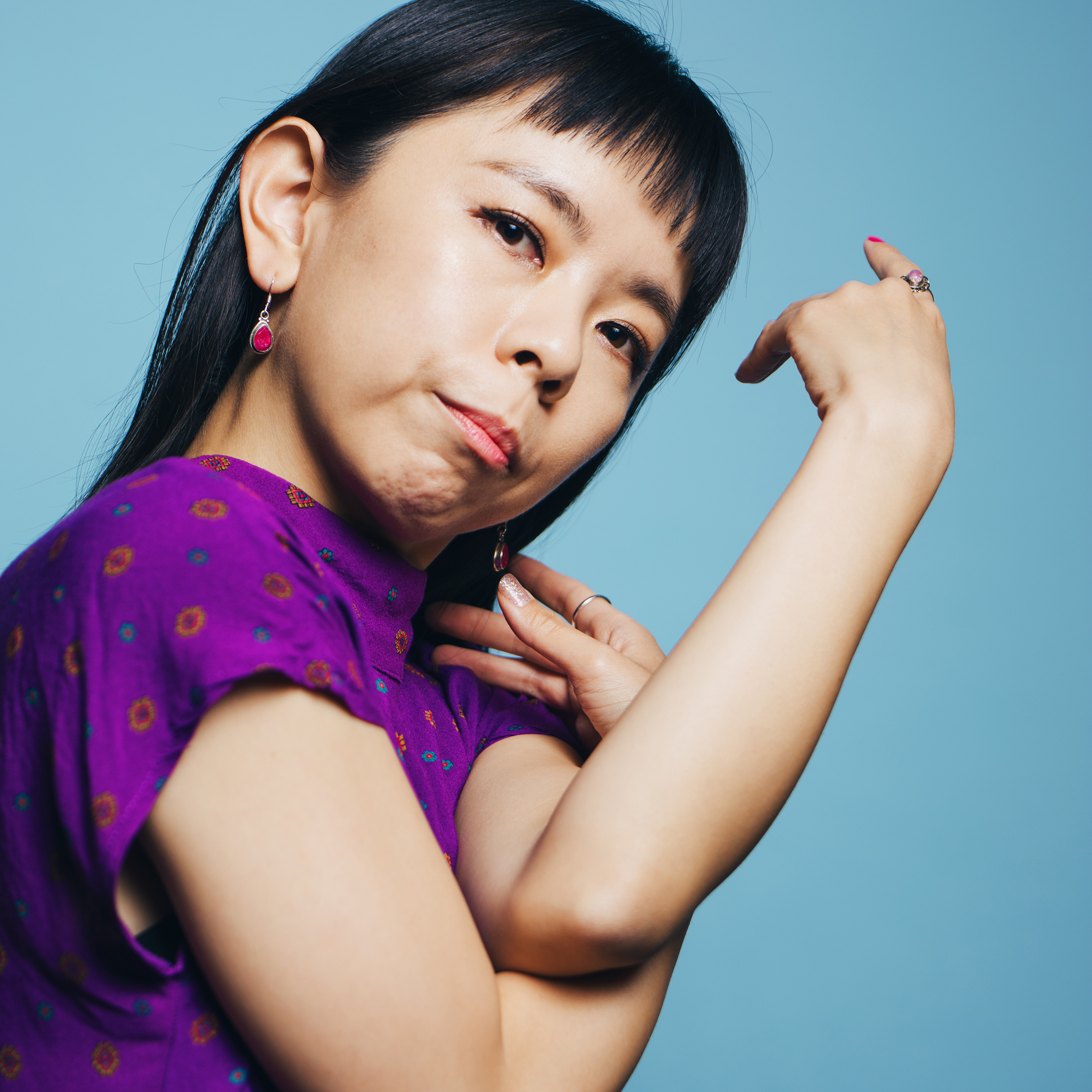 Portrait of Megumi Kokuba in a purple shirt, weaving her arms up close to her chest while she tilts her head to look at the camera