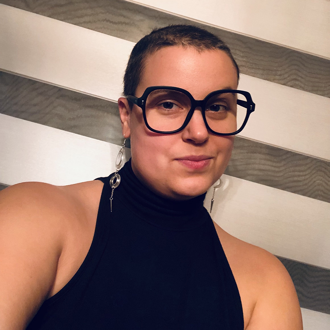 Kass Prus, a white skinned non-binary person looks directly at the camera with a small smirk. They have buzzed hair, thick black frame glasses, and are wearing a high necked black tank top and dangly clear and spiked earrings. The background is wide diagonal white and grey stripes