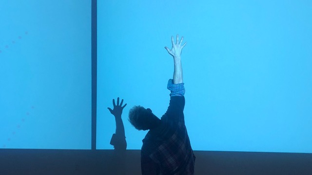 Performing, Mark Reinhart is turned towards the back, leaning his torso to the side while extending his arm straight in the air. He looks at the shadow of his arm that appears on the back wall.