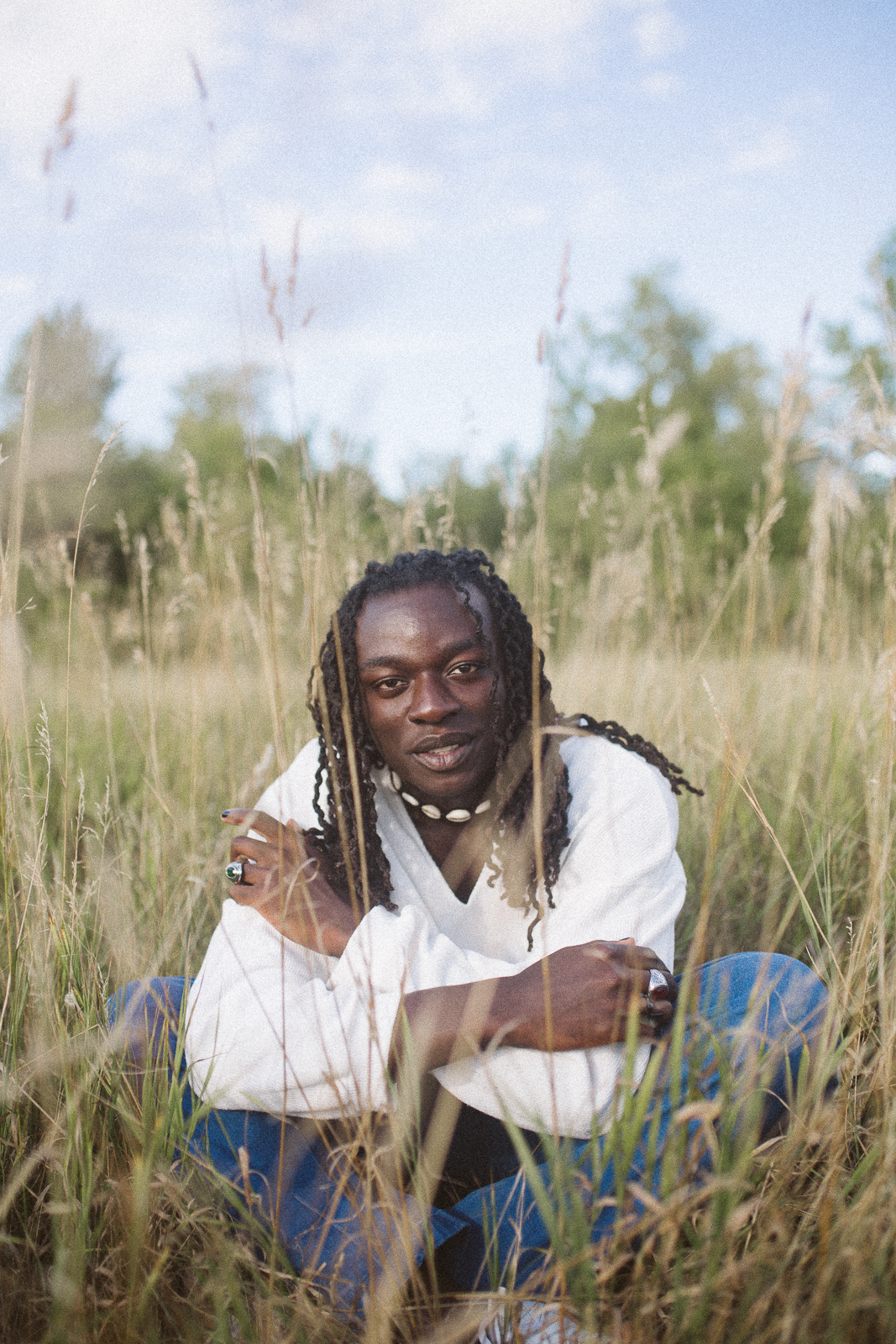 Headshot of Pilot Episode Choreographer Kwasi Obeng, dark skin male in white shirt and jeans surrounded in green field with trees and blue sky