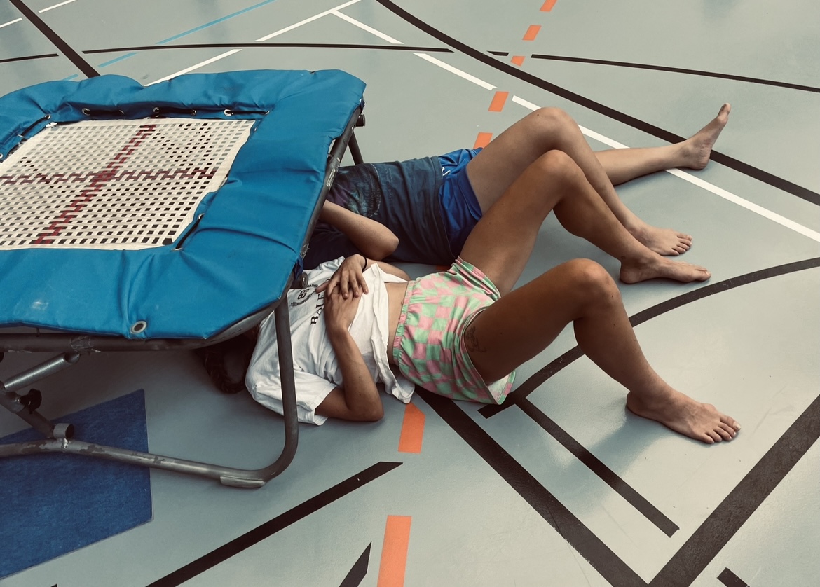 Two dancers lay on a studio floor with their heads underneath a small trampoline during dance class.
