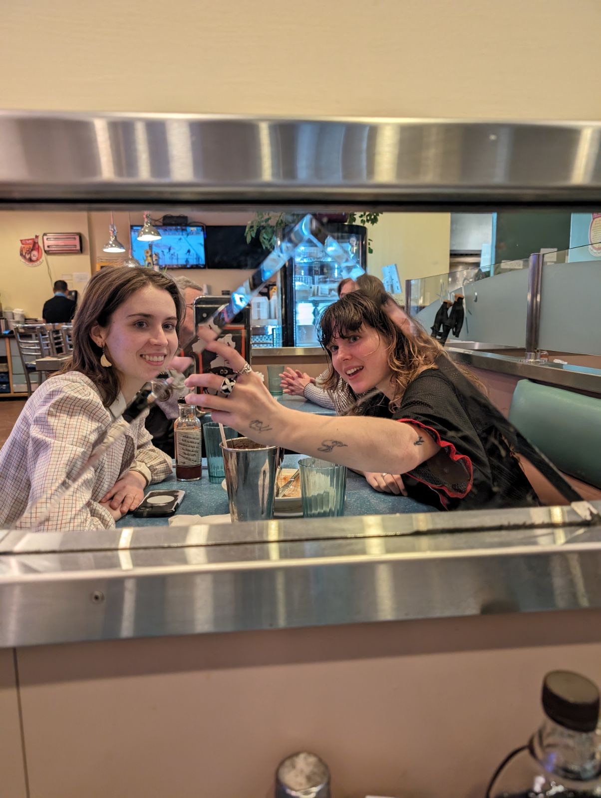 Two friends take a mirror selfie while sitting at a booth table.