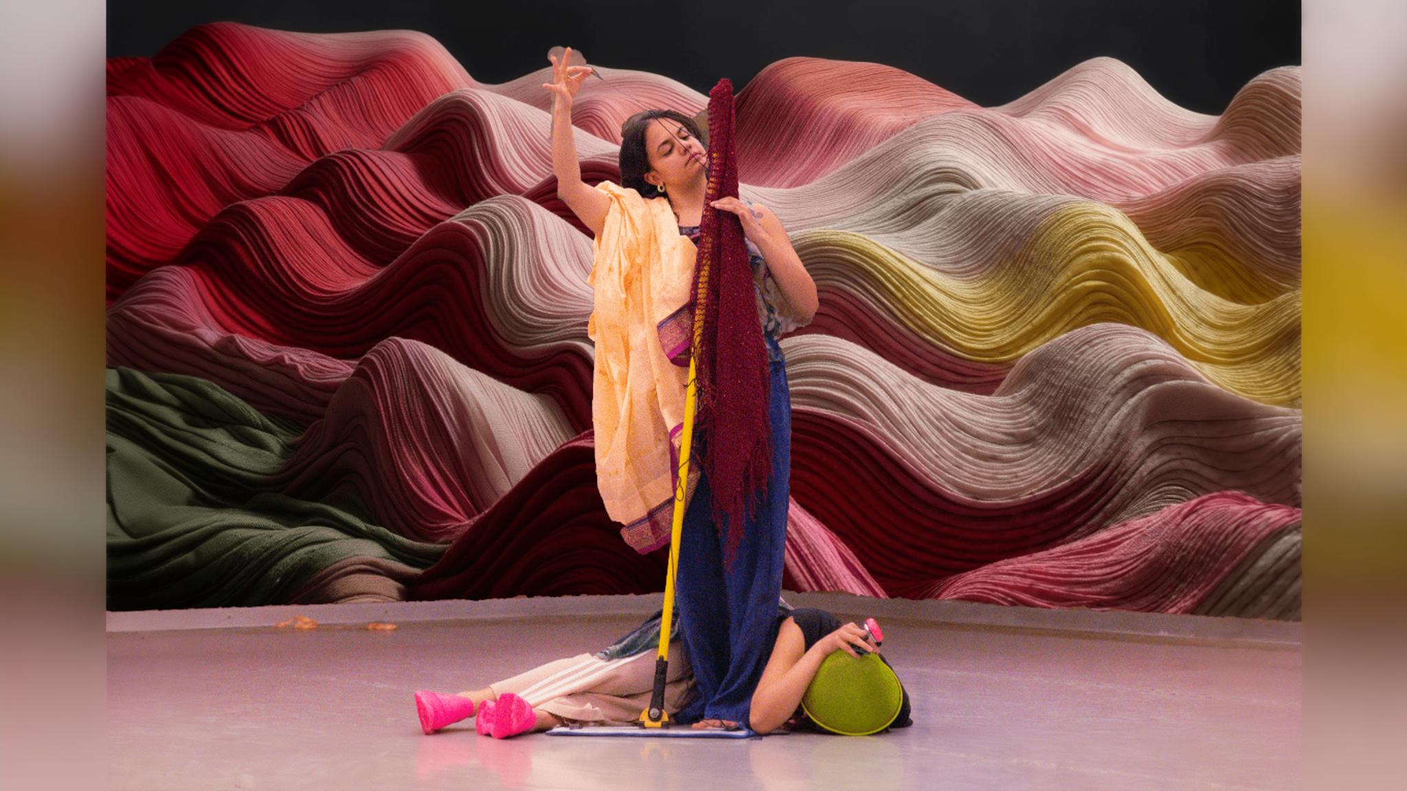 A performer stands in colorful attire on a stage, surrounded by swirling fabric patterns in shades of red, yellow, and green.